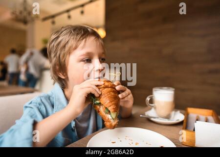 Happy cute boy eating fresh croissant, sitting at the table in city cafe, with paper cup of cocoa Stock Photo