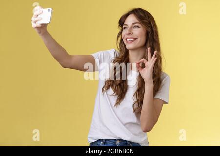 Stylish carefree urban confident curly-haired girl recording video blog followers extend arm hold smartphone taking selfie front camera show okay ok Stock Photo
