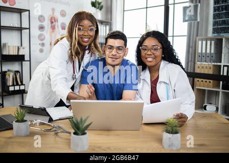 International team of three competent doctors in uniforms using modern gadgets for urgent meeting at conference room. Smiling colleagues analysing patient's medical record together. Stock Photo