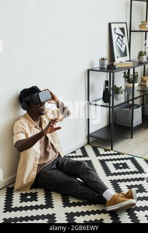 African excited man in virtual reality glasses playing video game while sitting on the floor in the room Stock Photo