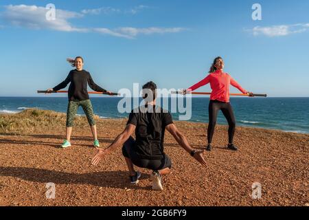 Middle aged man and woman trainer conduct fitness classes on the ocean shore. Using gymnastic sticks for stretching. Blue sky. Stock Photo