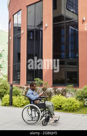 Disabled young black man in eyeglasses communicating online using smartphone while walking in wheelchair Stock Photo