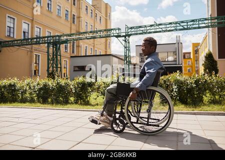 Sad pensive young black disabled man in glasses using wheelchair while walking down street alone Stock Photo