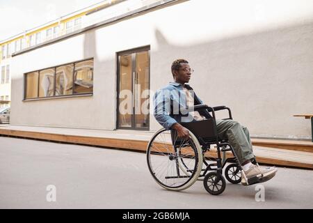 Serious handicapped young African-American man in eyeglasses using wheelchair while walking alone