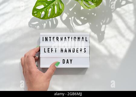 Text International Left Handers Day on light box in hand. Off white background with sunlight and shadows of monstera leaves. Flat lay, top view, hand Stock Photo