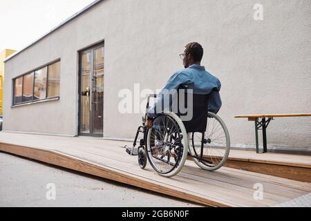 Rear view of handicapped African-American man in casual shirt using wheelchair ramp built by his house entrance Stock Photo
