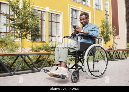 Content handsome young black man with disability sitting in wheelchair and browsing Internet on smartphone during stroll Stock Photo