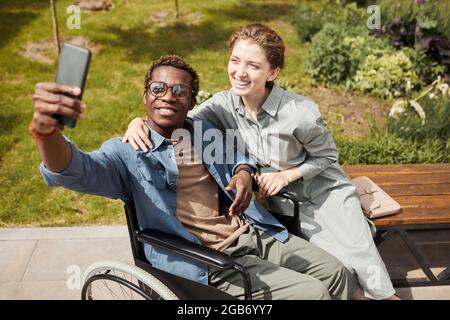 Happy young handicapped African-American man taking selfie with girlfriend in park while they spending leisure together Stock Photo