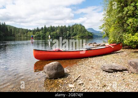 Canoeing in Scotland - Orange canoe on small beach and two paddle boarders passing by with Ben Lomond in the distance, Loch Ard, Trossachs, Scotland, Stock Photo