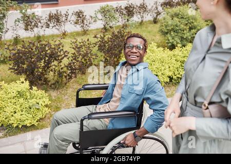Happy disabled young African-American man in glasses using manual wheelchair and talking to woman during date Stock Photo