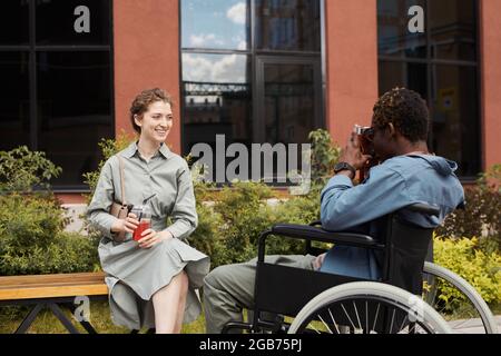 Disabled African-American photographer in wheelchair using camera while photographing young woman posing with summer beverage Stock Photo
