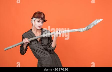 woman architect in protective helmet and boilersuit hold shovel on orange background, engineering Stock Photo