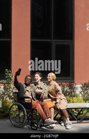 Positive young handicapped Black man in wheelchair using smartphone while taking selfie with friends on bench