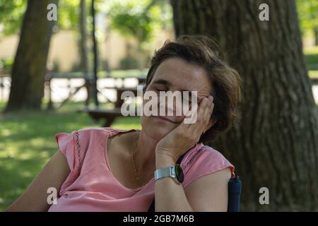 A middle-aged woman, tired from exhaustion, sits on a chair near a tree in a park on a sunny day. Stock Photo