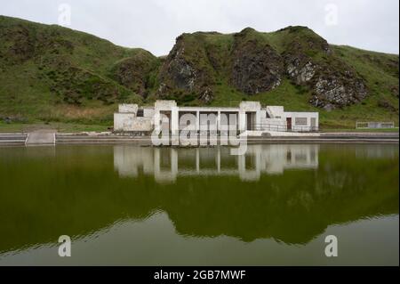 Abandoned art deco pavilion at Tarlair outdoor swimming pool, in Aberdeenshire. Reflection of building and surrounding hills in green water. Stock Photo
