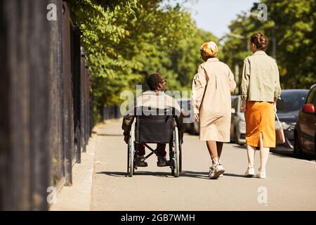 Rear view of handicapped black man in wheelchair walking with able-bodied friends and ommunicating with them Stock Photo