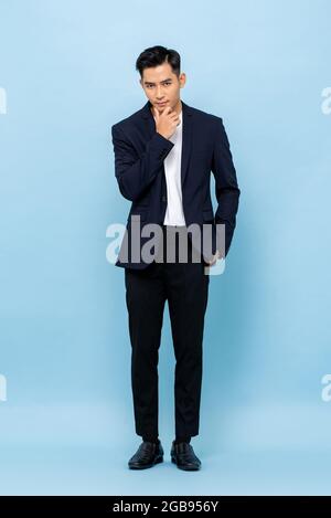 Full length portrait of young handsome Asian businessman standing and posing with hand touching chin on light blue studio background Stock Photo