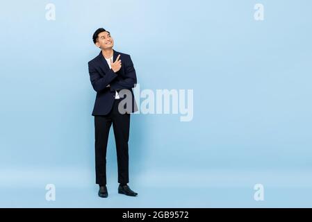Full length portrait of young handsome southeast Asian businessman looking up and pointing to copy space on light blue studio background Stock Photo