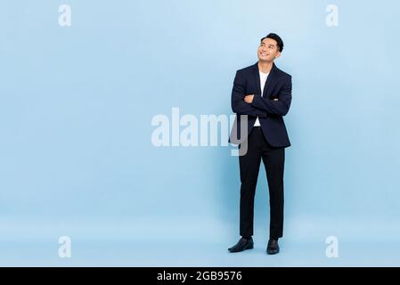 Full length portrait of young handsome southeast Asian businessman with arms crossed looking up to copy space on light blue studio background Stock Photo