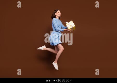 Full size profile photo of pretty brunette lady jump with present look empty space wear blue dress isolated on brown background Stock Photo