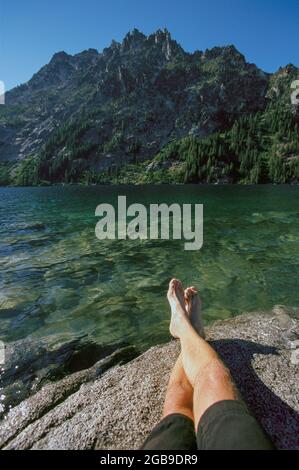 A mans legs seen from thigh down laying on a rock at the edge of a mountain lake. Stock Photo