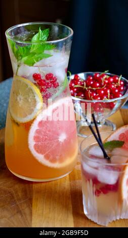 Grapefruit lemonade with mint, lemon, currants and ice in a decanter and a glass with black plastic tubes on a background of fruit. Cooling, refreshing, fruit, summer drink. Vertical close up Stock Photo