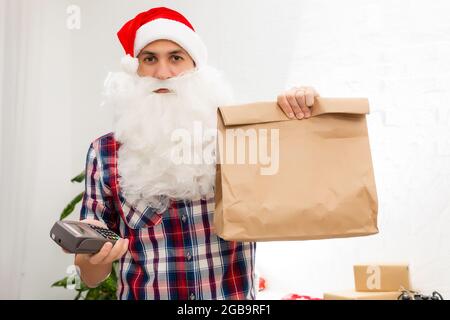 Positive smiling male courier in uniform and santa claus hat holding payment terminal and paper shopping bags, easy express order and delivery. Indoor Stock Photo