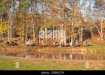 Group of people riding horses along a ranch trail beside trees and a pond in autumn, Pennsylvania, USA Stock Photo
