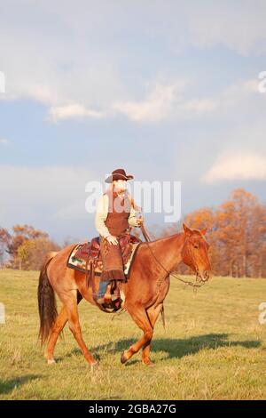 Woman riding a chestnut quarter horse through a ranch field in late afternoon sunlight Stock Photo