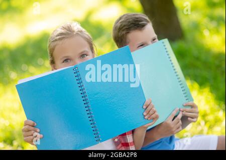 Girl and boy in playful mood hiding behind notebooks Stock Photo