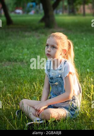 Little sad girl sitting in the park on the grass Stock Photo