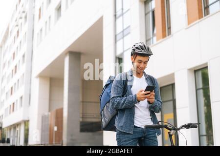 Medium shot of cheerful handsome male courier with thermo backpack standing with bicycle in city street and using navigation app on phone. Stock Photo