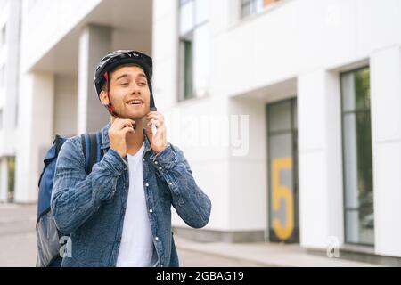 Front view of joyful young courier male with thermo backpack putting on helmet on head before to deliver an order to customer by bicycle. Stock Photo