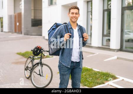 Portrait of smiling handsome young delivery man with large thermo backpack standing on city street. Stock Photo