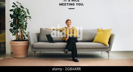 Smiling businesswoman sitting on sofa in office lobby. Cheerful entrepreneur on sofa. Stock Photo