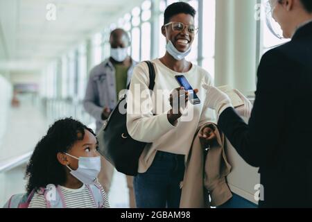 Woman traveler with daughter in face masks showing digital boarding pass at departure gate of airport. Airport attendant checking digital air ticket o Stock Photo
