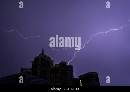 Colorful flash of lightning against the dark night sky above the roof of a building with a spire during thunderstorm and rain Stock Photo
