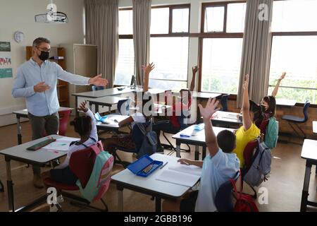 Group of diverse students wearing face masks raising their hands in the class at school Stock Photo