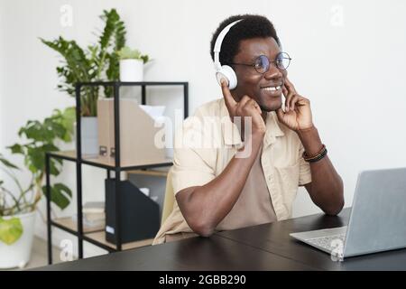 African smiling man putting on headphones and talking online using laptop while sitting at the table at office Stock Photo