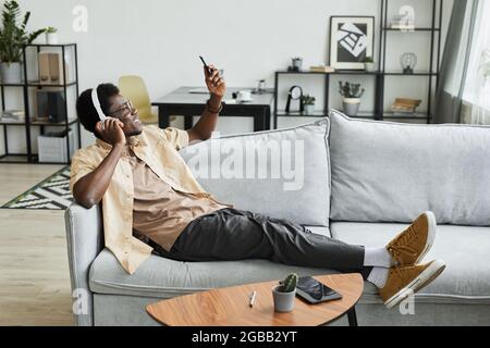 African smiling man in headphones looking at mobile phone and talking on video online while lying on sofa at home Stock Photo