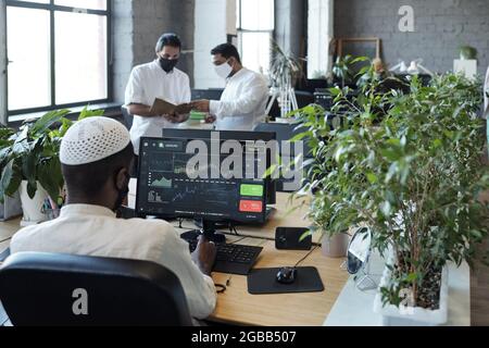 Rear view of young Muslim businessman sitting in front of computer monitor and his two colleagues in office Stock Photo