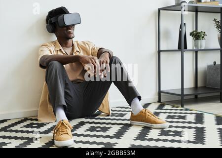 African young man sitting on the floor in the room in virtual reality glasses watching something online and smiling Stock Photo