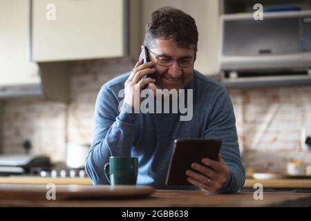 Smiling caucasian man in kitchen drinking coffee, using tablet and smartphone Stock Photo
