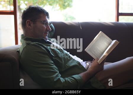 Smiling caucasian man lying on sofa, reading book and relaxing Stock Photo