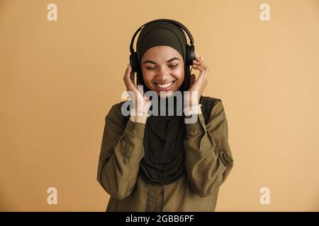 Black muslim woman in hijab listening music with headphones isolated over beige background Stock Photo