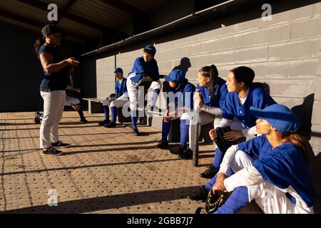 Diverse group of female baseball players sitting on bench, listening to female coach before game Stock Photo