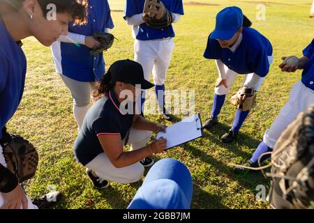 Diverse group of female baseball players in huddle around squatting female coach with clipboard Stock Photo
