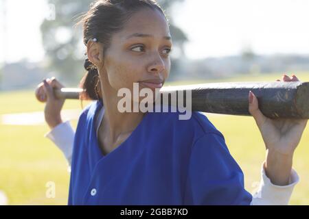 Mixed race female baseball player holding baseball bat on shoulders looking away on baseball field Stock Photo