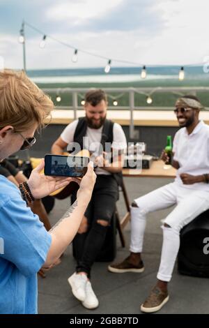 Young man shooting video on smartphone of his happy friends enjoying rooftop party Stock Photo