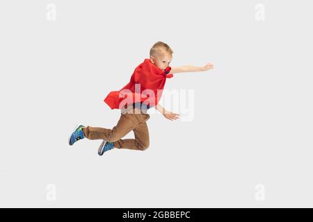 Portrait of little blond boy, pupil flying like super hero in red cape isolated on white studio background. Copyspace for ad. Childhood, education Stock Photo